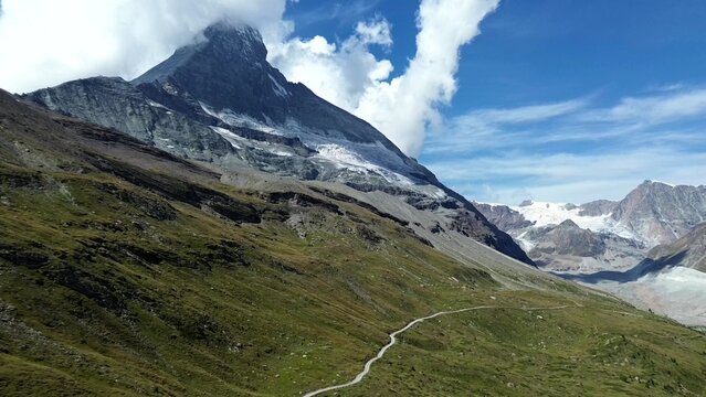 Aerial view of the Matterhorn Glacier with rugged mountain slopes, a winding hiking trail, and snow-capped peaks under a blue sky in Zermatt, Valais, Switzerland.