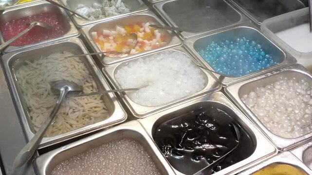 Close-up of colorful bubble tea and dessert toppings at a Chinese night market food stall, including tapioca pearls, jellies, and syrups in stainless steel trays