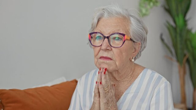 Elderly woman with hands clasped in prayer, red nails and glasses seated on a couch in a building; contemplation.