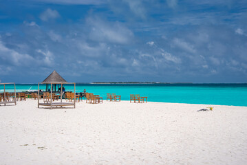 Fototapeta premium White sand stretches to turquoise water with lounge chairs and a thatched pavilion in the Maldives. Soft daylight and wide view suggest a quiet resort setting.