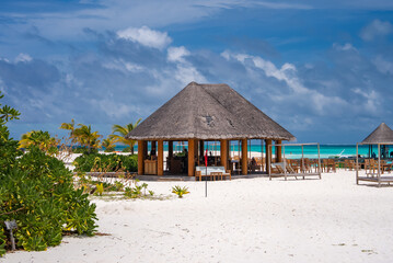 Fototapeta premium A thatched pavilion on white sand sits by turquoise water in the Maldives. Open sides reveal a bar and dining area, with cabanas and palms under midday light.