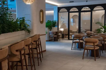 Softly lit lounge interior in the Maldives with tan leather barstools, white stone counter, rounded wooden chairs, arched glass partitions, plants, and pendant. © Aerial Film Studio