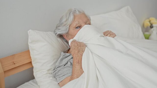 Woman in bed pulls white duvet over face while gripping sheet beside wooden headboard in a building bedroom; vulnerability aging.