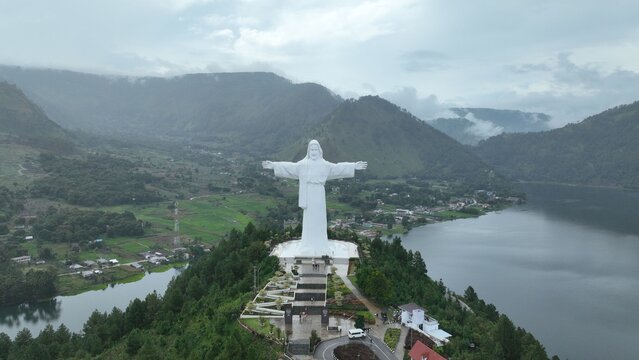 Aerial view of the Sibea Bea statue standing on a hill overlooking Lake Toba with lush green landscapes, winding roads, and cloudy skies in Samosir Regency, North Sumatra, Indonesia.