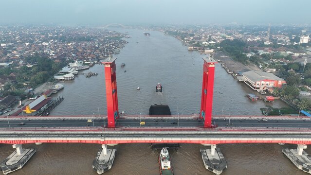 Aerial view of Jembatan Ampera spanning the Musi River with a barge being towed underneath, surrounded by the urban landscape of Kecamatan Seberang Ulu I, Sumatera Selatan, Indonesia.