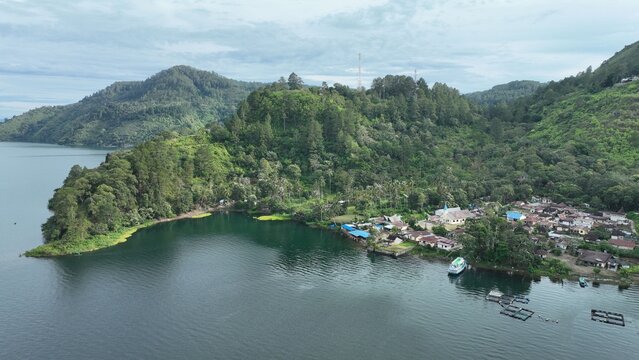 Aerial view of a village nestled along the lush green shoreline of a lake with fish farming cages floating in the water, Kecamatan Seberang Ulu I, Sumatera Selatan, Indonesia.
