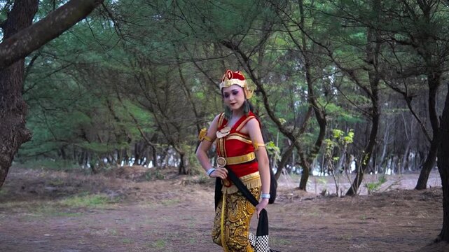 Traditional performer in ornate red and gold costume walks and gestures through shaded wooded park, captured near end of clip with soft daylight and graceful