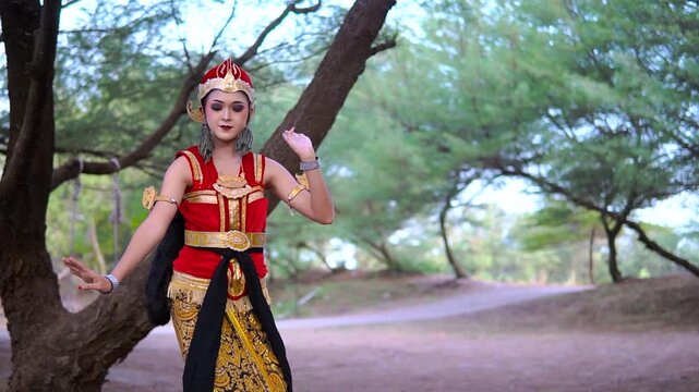 Female performer in ornate red and gold costume holds graceful dance pose beside shaded tree-lined path, calm expression, soft natural light
