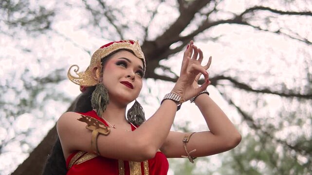 Woman in ornate red costume and headpiece performs poised hand movements outdoors beneath bare tree branches, with calm focus and blurred natural background