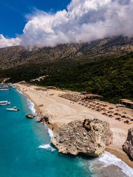 Aerial view of the coastline in Sarande, featuring turquoise waters, a pebble beach with rows of sun loungers, large coastal rocks, and lush green hills in Sarande, Vlore County, Albania.