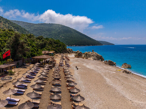Aerial view of a beach with rows of straw umbrellas, sun loungers, and the Albanian flag, set against hills and the turquoise Ionian Sea in Sarande, Vlore County, Albania.