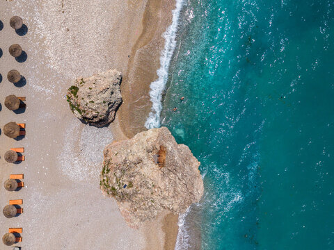 Aerial view of large rocks on a sandy beach with rows of umbrellas and lounge chairs along the turquoise Ionian Sea in Sarande, Vlore County, Albania.