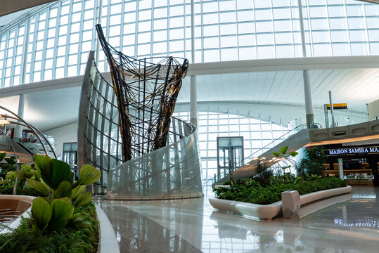 Abu Dhabi airport terminal interior shows twisting metal rods rising from a curved glass base, floor to ceiling windows, tropical planters, polished stone floors, and MAISON SAMIRA signage.