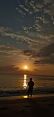 Fototapeta premium A Solitary Man Standing on the Beach Shore Watching the Beautiful Golden Sunset Over the Ocean Waves at Dusk