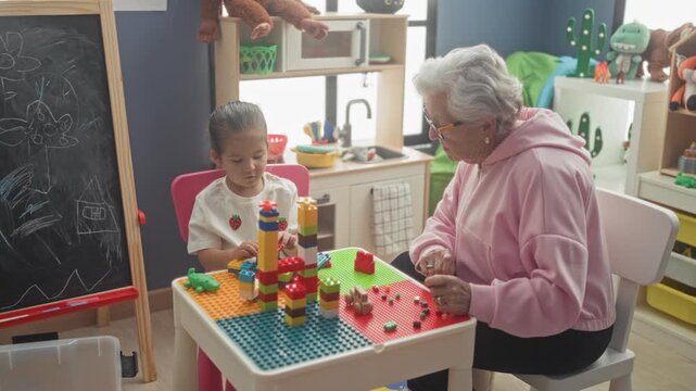 Woman helps little girl stack colorful toy blocks at a small play table with chalkboard and kitchenette visible in a building; intergenerational bonding.