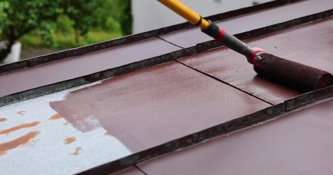 man painting tin metal roof with roller and brown paint. house roofing restoration