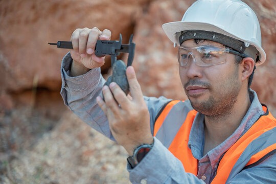 Professional Asian male geologist wearing safety gear uses a caliper to precisely measure a rock specimen during fieldwork at a rugged mining quarry for industrial environmental evaluation.
