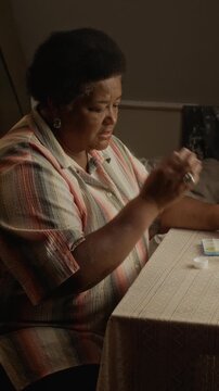 Senior African American woman taking daily medication from pill organizer and drinking water. Ideal for senior healthcare, pharmaceutical, or medical insurance advertising, vertical