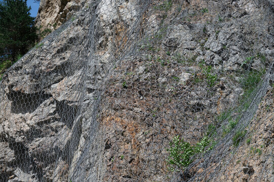 Close-up of industrial steel wire mesh drapery system installed on a dangerous rocky cliff to prevent rockfalls and landslides.