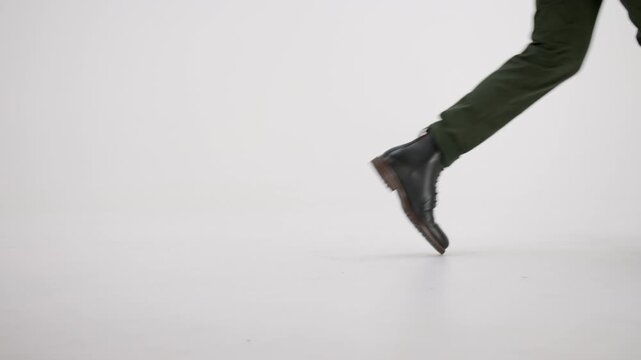 Lower body view of a man walking from left to right. He is wearing stylish black leather boots and green pants, isolated on a white studio background