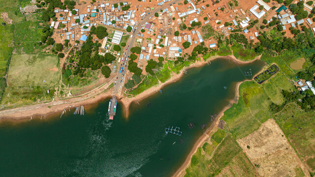 Aerial view of a ferry docked at the riverbank near village buildings, lush green vegetation, and calm water in Kwahu Afram Plains South, Eastern Region, Ghana.