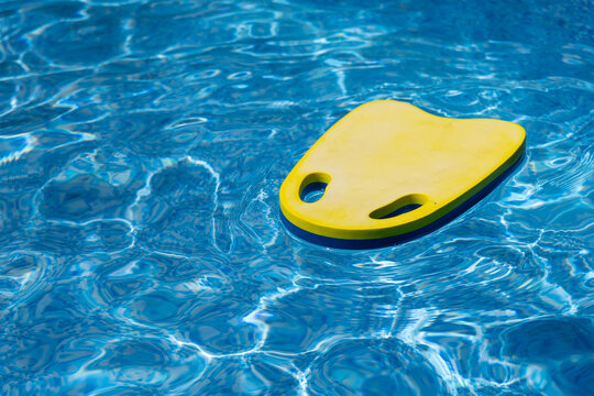 A swimming kickboard floats on the surface of a swimming pool. The scene shows clear blue water and a calm summer atmosphere, highlighting basic swim training equipment used for learning and practice.