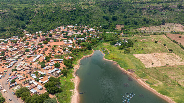 Aerial view of a village along a riverbank with small houses, dirt roads, lush hills, and fish farming cages in Kwahu Afram Plains South, Eastern Region, Ghana.