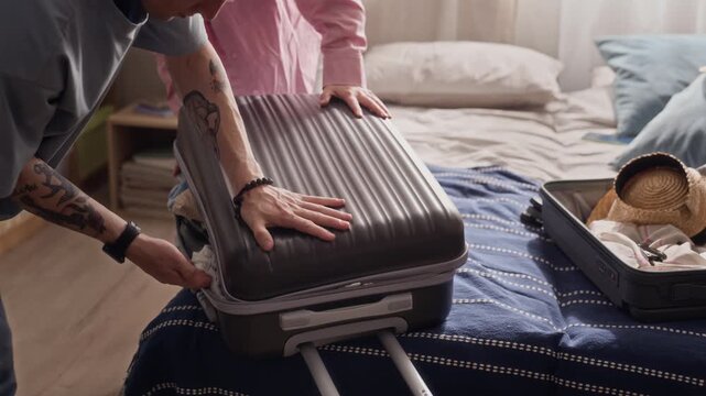 Handheld shot of Caucasian young couple struggling to close overpacked suitcase on bed, woman kneeling on luggage to help man zip it. Ideal for travel agency commercials or luggage brand ads