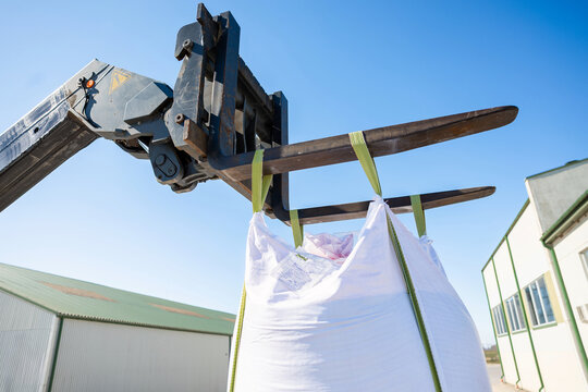 A telehandler with extended forks lifts a white bulk bag by green straps at a warehouse gate, seen from a low angle beside corrugated buildings under bright midday light.