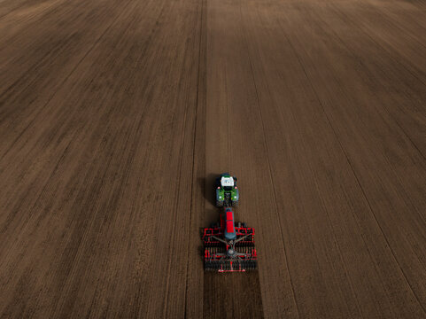 Aerial view shows a single green tractor pulling a red harrow across a vast tilled field with parallel furrows, soft even daylight, minimal composition, no structures