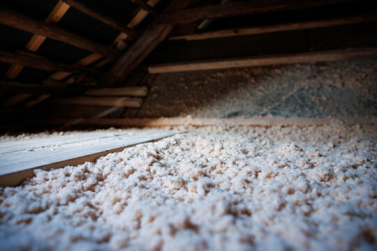 Inside an older wooden house in Riga, a low angle view shows thick cellulose insulation on the attic floor, exposed beams, a narrow plank path, and warm, low lighting.