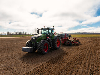 Fototapeta premium A large green Fendt tractor with red wheels tows a multi row seed drill across freshly tilled soil, cutting straight furrows under low clouds during midday planting.
