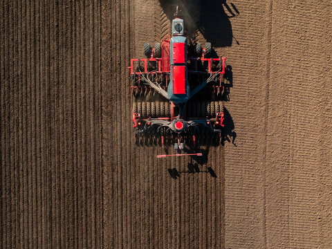 Aerial view shows a red tractor and seeder carving parallel furrows in dry tilled soil. Midday light casts crisp shadows, highlighting mechanized planting precision.