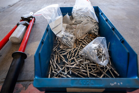 A blue bin holds many metal blind rivets, some in opened bags, beside a red hand riveter on a concrete floor in a workshop. Natural light and shallow depth of field show detail.