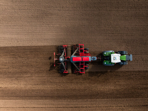 Aerial top down view shows a green and white tractor towing a red seed drill across brown soil, carving parallel furrows with multiple coulters and a hopper system.