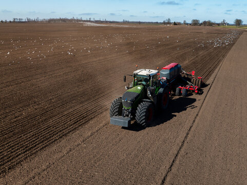 Aerial view of a Fendt tractor towing a red seed drill on tilled rows, white birds scatter over an expansive field under cool daylight with thin clouds.