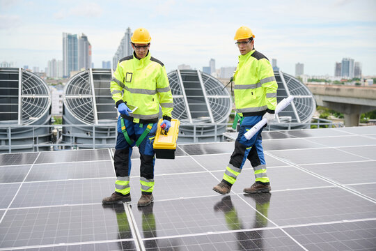 Technicians inspecting a solar panel array on a rooftop, dressed in safety gear. One is holding a blueprint and the other a toolbox. They appear focused on their work