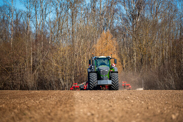 Obraz premium A large green tractor with dual front tires pulls red tillage gear across fresh furrows, dust rising. Bare deciduous trees stand beyond, cold light and low angle frame power.