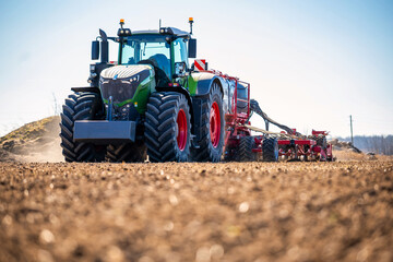 Fototapeta premium Modern Fendt tractor with oversized tires tows a red multi row seeder across tilled soil, dust rises, GPS guided planting in spring, low angle view shows tread and hoses.