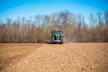 Obraz premium A green tractor with dual lights pulls red tillage toward the viewer on a freshly plowed field, dust rising, bare deciduous treeline in bright daylight, cool season.