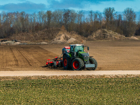 A large green tractor with red wheels tows a seed drill across tilled soil, leaving neat parallel furrows. Soft daylight and cool clouds light a gravel path and bare trees.