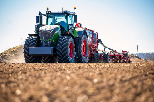 Modern Fendt tractor with oversized tires tows a red multi row seeder across tilled soil, dust rises, GPS guided planting in spring, low angle view shows tread and hoses.