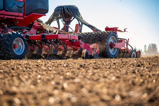 A red air seeder with discs, packer wheels, and hoses moves across tilled soil, dry dust rises, hydraulic lines show, daylight is crisp, rows form in a broad field.