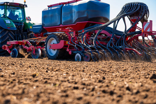 A green tractor tows a red air seeder with hoses and openers, cutting parallel furrows as dust rises from prepared soil. Side light defines seed tanks and lines.