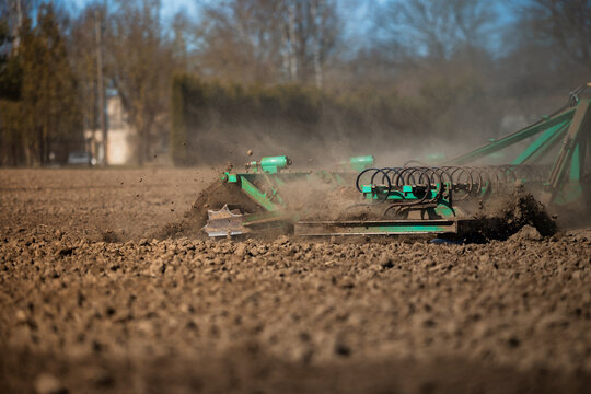 Side view shows a green cultivator tearing dry, cloddy soil, dust rising, springs vibrating, bare trees and distant structures blur, early spring farm work underway.