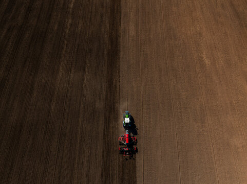 Aerial view of a green tractor pulling a red plow, carving parallel furrows that split dark tilled soil and light soil, dust trailing under strong sunlight.