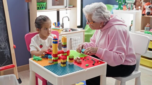 Senior woman stacks colorful building blocks and hands a block to little girl at a playtable in building; intergenerational play warmth.