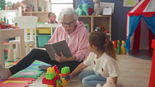 Elderly woman reading book to little girl, hand on page, seated on floor toys and tent visible in building playroom; intergenerational bonding warmth.