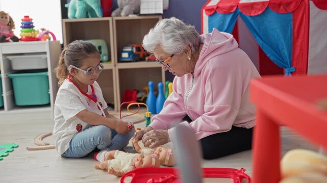Elderly woman and little girl sit on floor and tend to baby doll, hands holding doll and pointing at toy inside playroom building; tenderness bonding care.