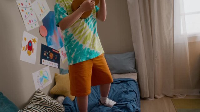 Caucasian boy with Down syndrome standing on bed and playing ukulele in bedroom. Clip perfect for advertisements promoting social diversity, inclusion, and special education programs